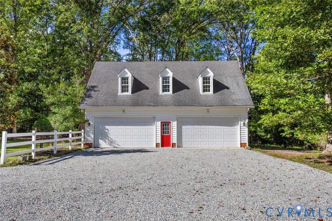 Two-car garage with white paneled doors, three dormer windows, a vibrant red entry door, and a gravel driveway