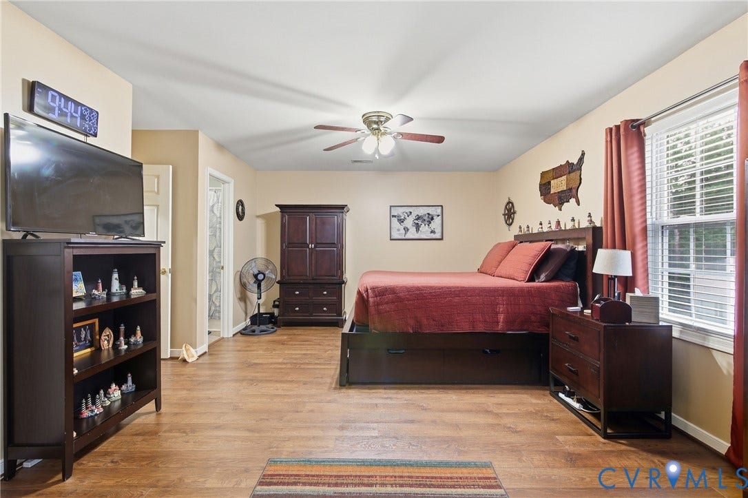 10624 Hamlin Drive, Hamlin Estates, Chester, VA 23831 Spacious primary bedroom suite featuring hardwood flooring, a ceiling fan with integrated lighting, ample natural light from dual windows with red curtain treatments, dark wood bedroom furniture, dark wood cabinetry with shelving, a wall-mounted flat-screen television, and an attached walk-in closet accessible via double wooden doors