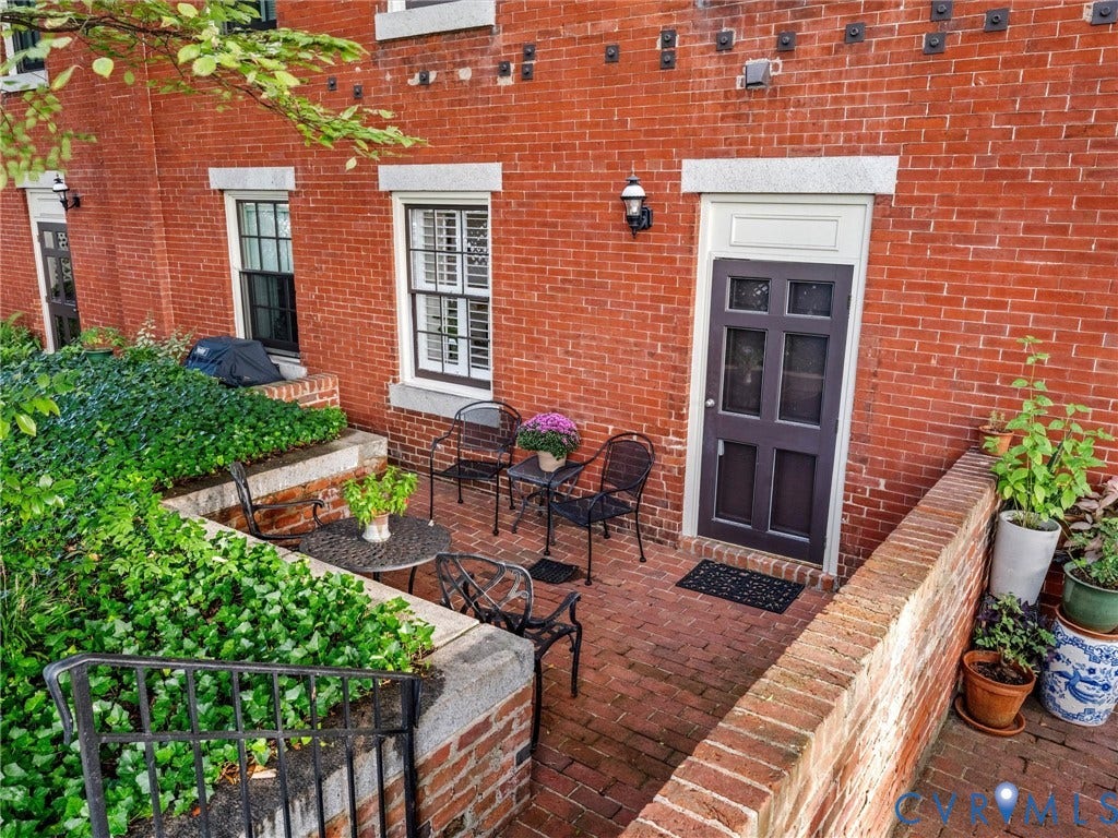 Brick exterior featuring a paneled entry door, shuttered windows, a brick patio, and tiered landscaping