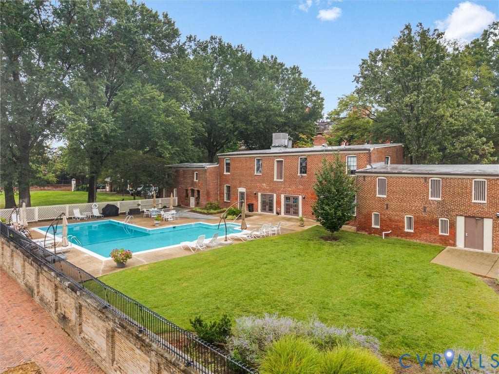 Brick exterior structure with multiple windows, an in-ground swimming pool, a surrounding patio, lush green lawn, and mature trees
