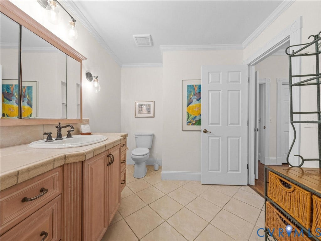Light-toned tile flooring, wood-finish vanity with countertop, framed mirror, and crown molding