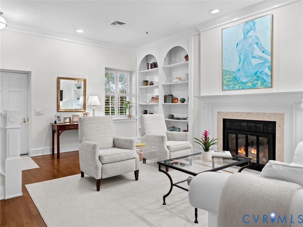 Living area featuring a fireplace with a stone surround, built-in arched shelving, crown molding, wood-finish flooring, and a window with plantation shutters