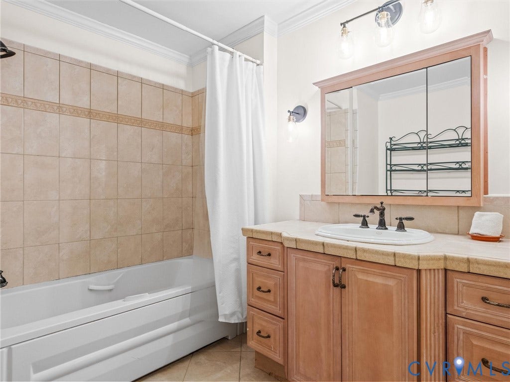 Tiled bathtub surround with a decorative accent band, white built-in tub, wood-finish vanity with an integrated sink, tiled countertop, and a mirrored medicine cabinet