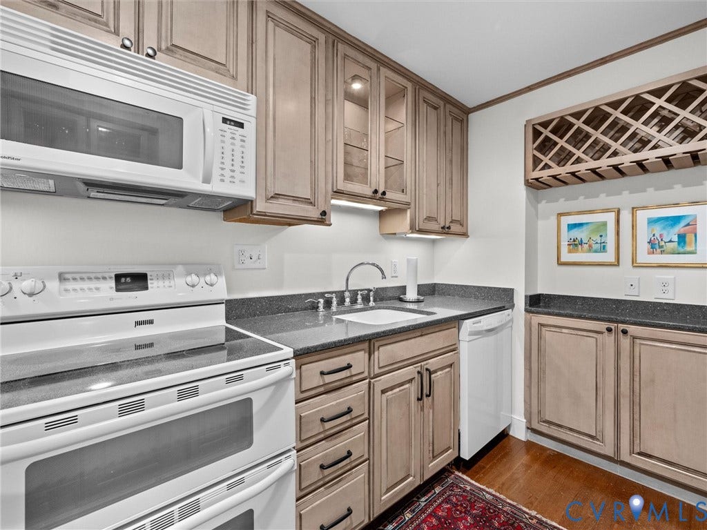 Wood-finish flooring, light wood-finish cabinetry with dark hardware, granite-style countertops, white double oven, and a built-in wine rack