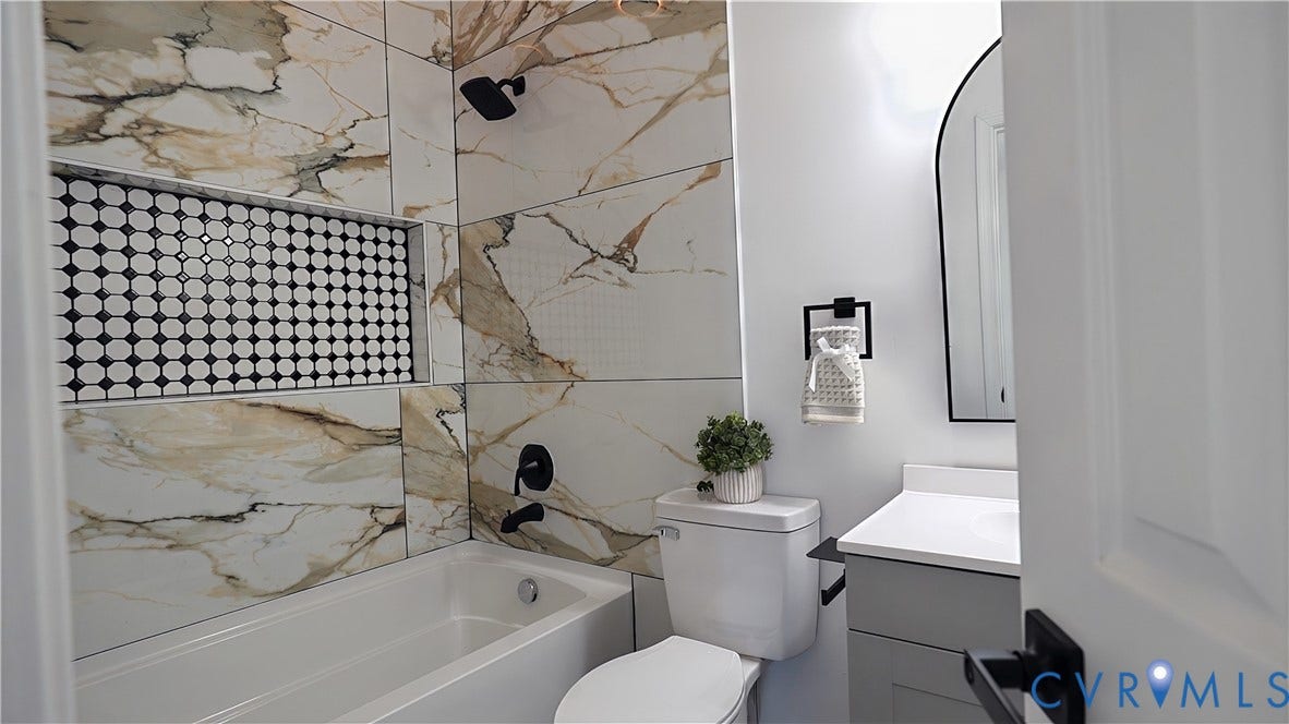 326 Mars Street, Petersburg, VA 23803 Bathroom featuring a built-in tub with matte black fixtures, an arched vanity mirror, a single vanity with a white countertop, and large format wall tiles with prominent veining