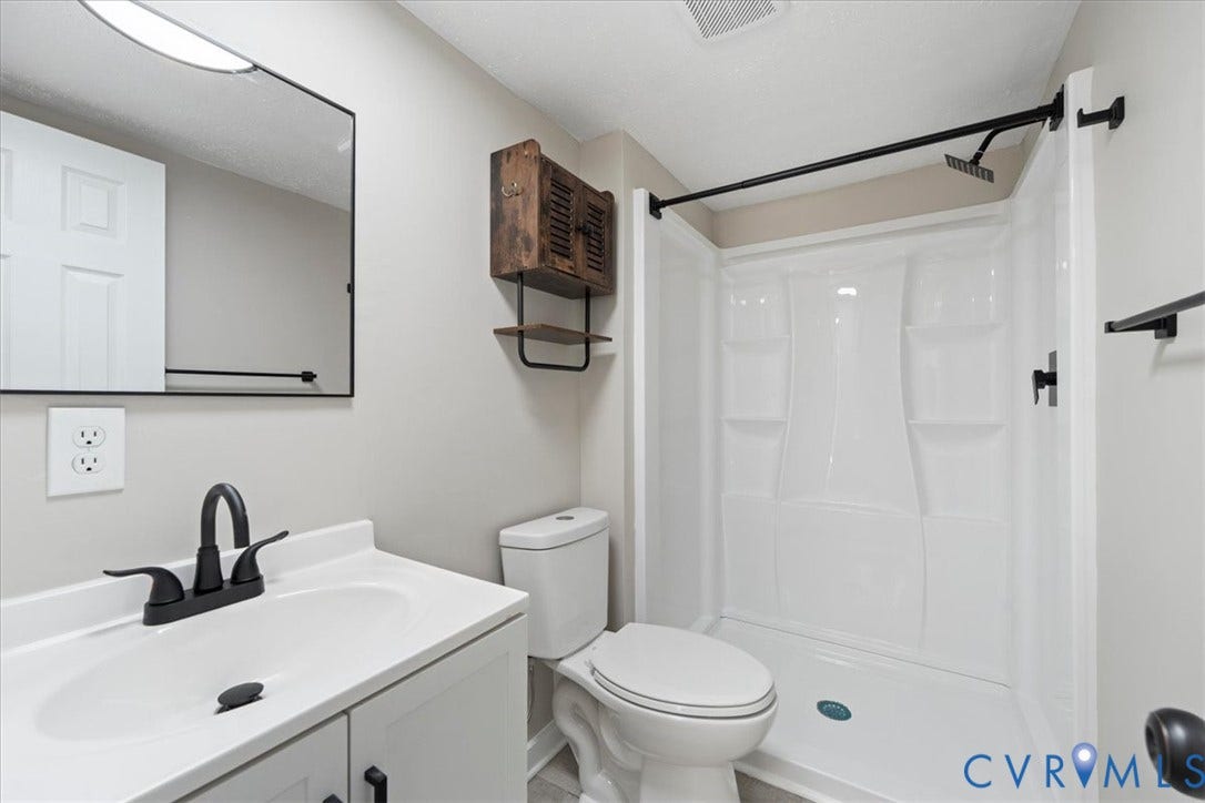 115 Mount Airy Lane, Cumberland, VA 23040 Modern bathroom featuring a white vanity with an integrated rectangular sink, matte black faucet and hardware, large framed mirror with black trim, white subway tile shower surround with glass enclosure, white toilet, and woven storage basket