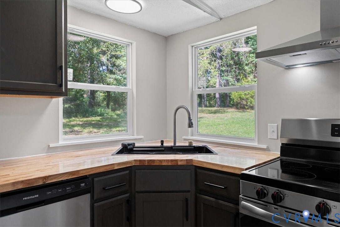 115 Mount Airy Lane, Cumberland, VA 23040 Modern kitchen featuring a corner sink with matte black gooseneck faucet, wood-finish countertops, black cabinetry with hardware, stainless steel appliances including a gas range, and abundant natural light from oversized windows overlooking landscaped grounds