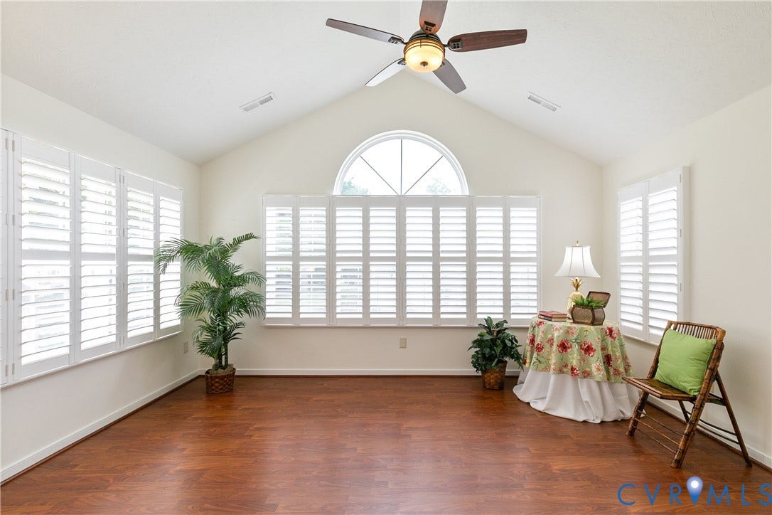 Vaulted ceiling with a ceiling fan, wood-finish flooring, multiple windows with white plantation shutters, and a prominent arched window