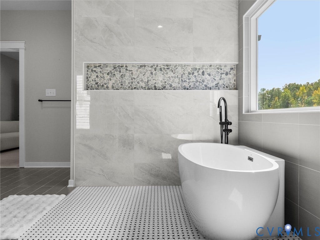 Freestanding soaking tub with a floor-mounted matte black faucet, surrounded by large format tile walls with a pebble mosaic accent