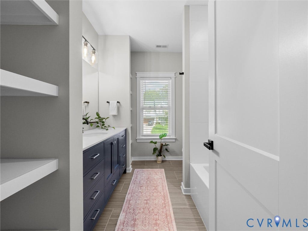 Floating shelving, a double vanity with ample storage, a window with horizontal blinds, and a built-in bathtub with subway tile surround