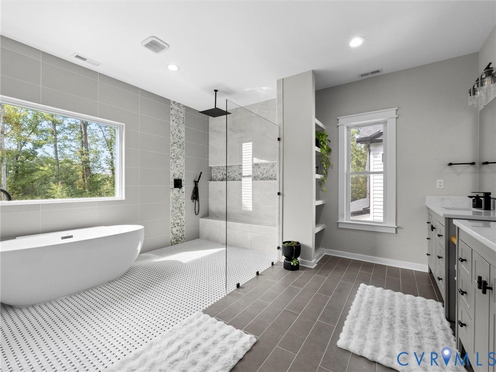 Spa-inspired primary bathroom featuring a freestanding soaking tub, walk-in shower with frameless glass enclosure and rainfall showerhead, dual vanity with white cabinetry and quartz countertops, heated tile flooring in a herringbone pattern, and abundant natural light from large windows overlooking mature landscaping