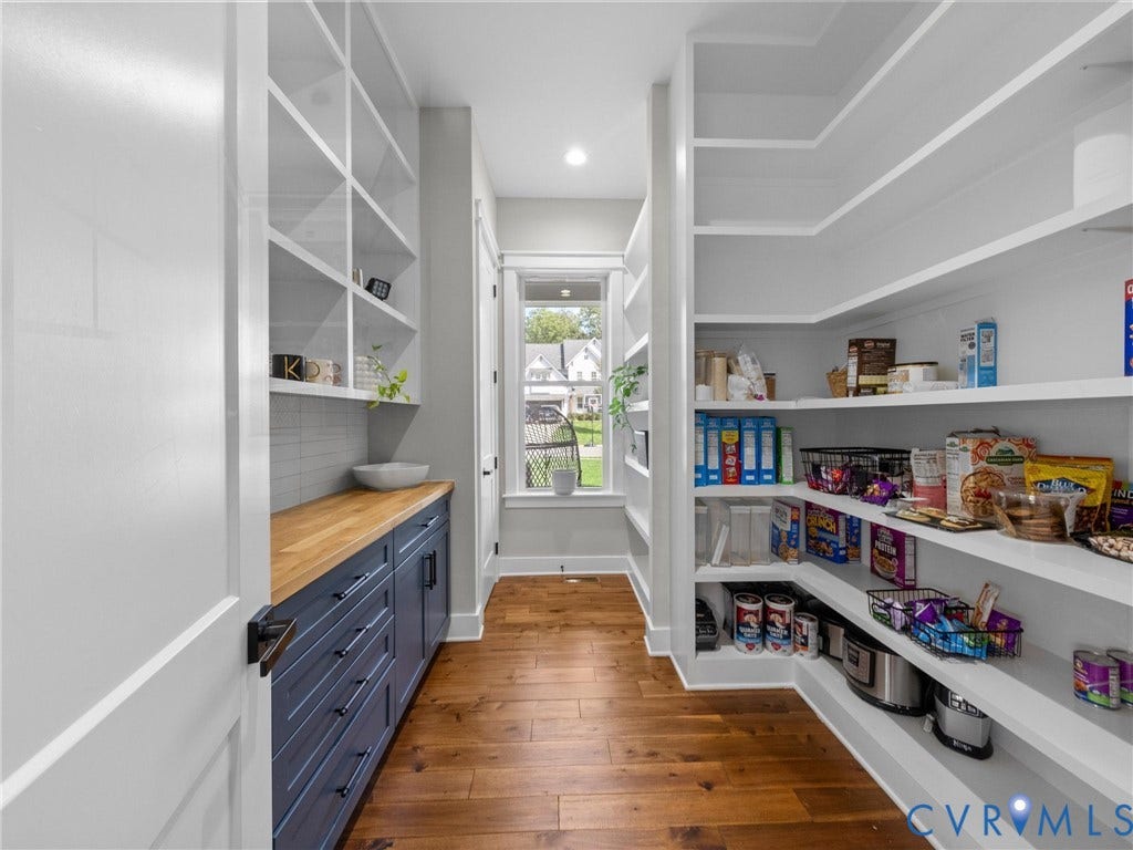 Well-appointed pantry featuring wood-finish flooring, extensive built-in shelving, blue cabinetry with a wood countertop, and a tiled backsplash