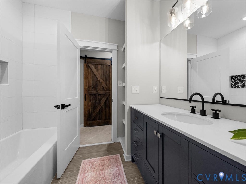 Bathroom featuring a built-in tub with white subway tile surround, dark wood-finish sliding barn door, dark cabinetry with white countertop, and contemporary vanity light fixtures