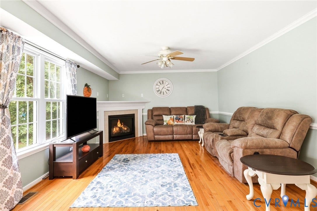 Hardwood flooring, corner fireplace with white mantel, ceiling fan, and large windows with exterior views