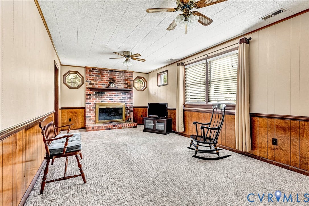Brick fireplace with a brass-finish insert, wood-finish wainscoting, carpeted flooring, and two ceiling fans