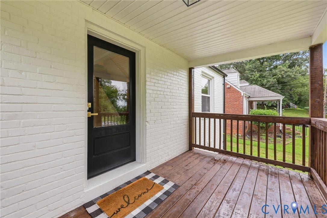Covered porch featuring white-painted brick, a wood-finish floor, wood railings, and a black entry door with glass panel