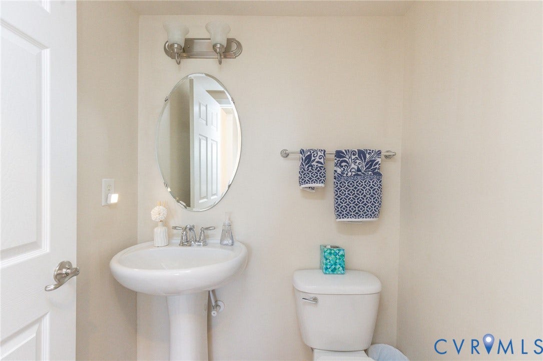 Pedestal sink, oval mirror, chrome wall-mounted light fixture, white toilet, and chrome towel bar