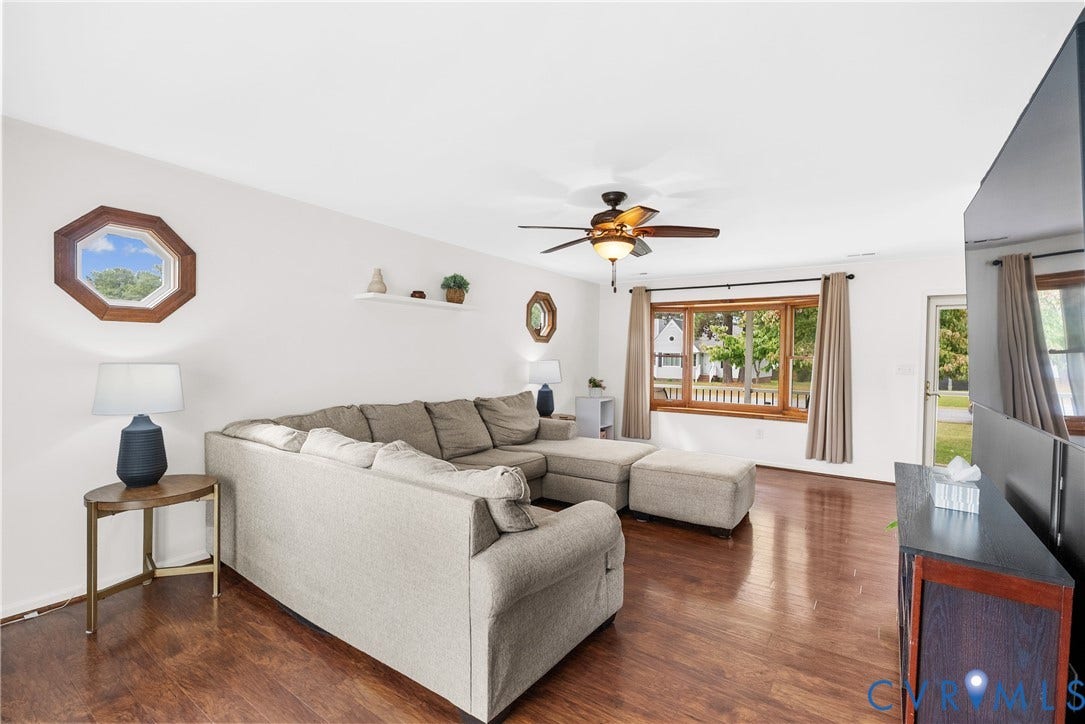 Wood-finish flooring throughout, ceiling fan with integrated lighting, large picture window with wood trim, and an octagonal accent window