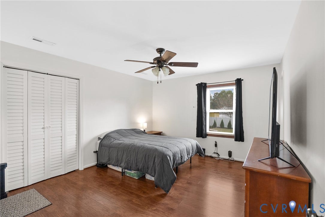 Wood-finish flooring throughout, white walls, louvered bi-fold closet doors, ceiling fan, and a window providing natural light