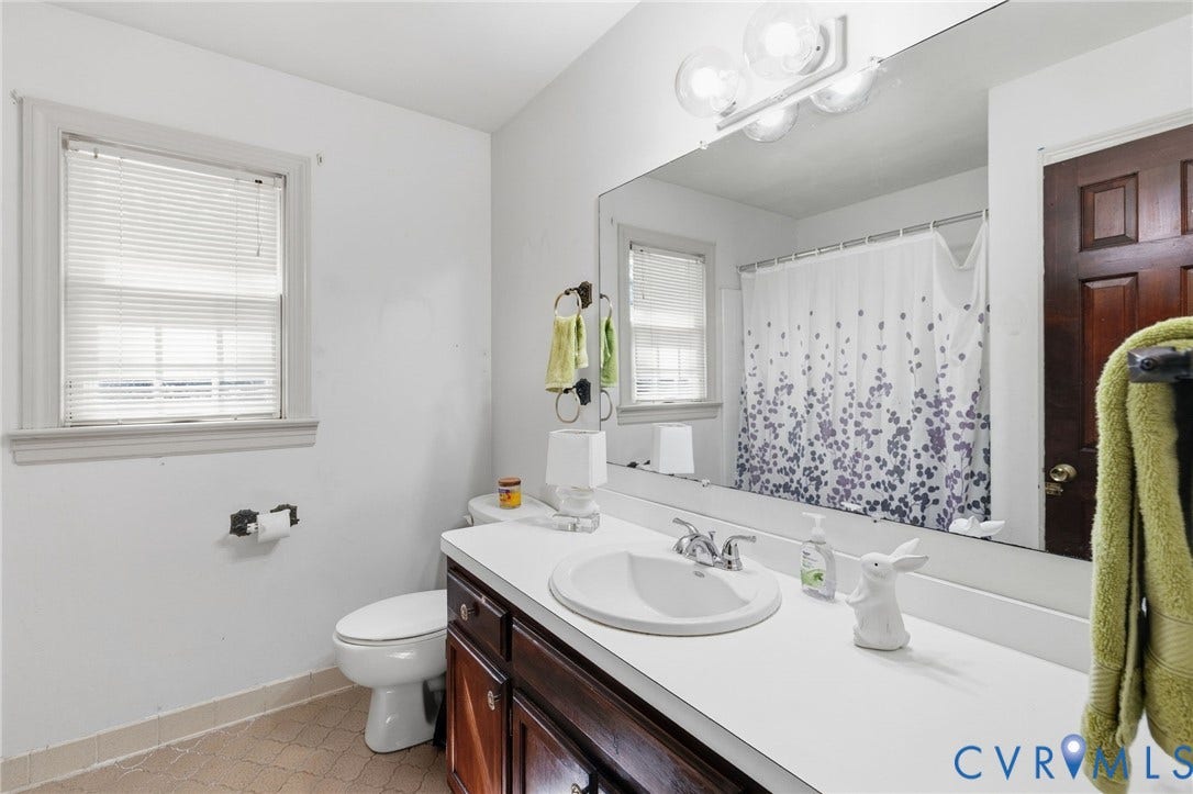 Bathroom featuring a single window with blinds, a vanity with a white countertop and dark wood-finish cabinetry, an oval undermount sink, and a large wall-mounted mirror