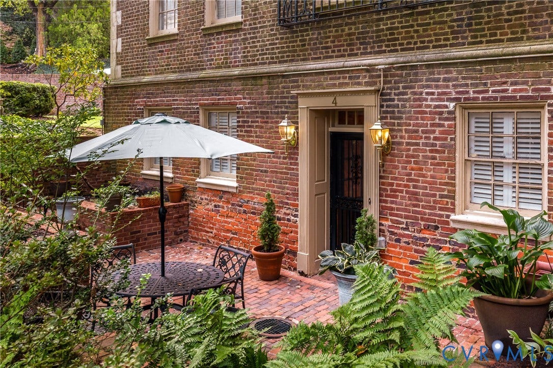 5621 Cary Street Road, #U4, Wilton, Richmond, VA 23226 Classic brick exterior featuring a multi-pane entry door with metal screen, multi-light windows with louvered shutters, traditional wall-mounted carriage lanterns, and a herringbone-patterned brick patio