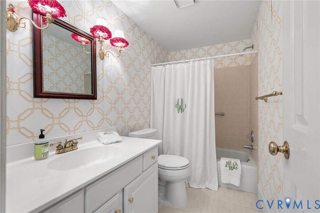 5621 Cary Street Road, #U4, Wilton, Richmond, VA 23226 Bathroom featuring a vanity with a white solid surface countertop, brass-finish faucet hardware, patterned wallcovering, dual brass-finish wall sconces, and a tiled shower surround