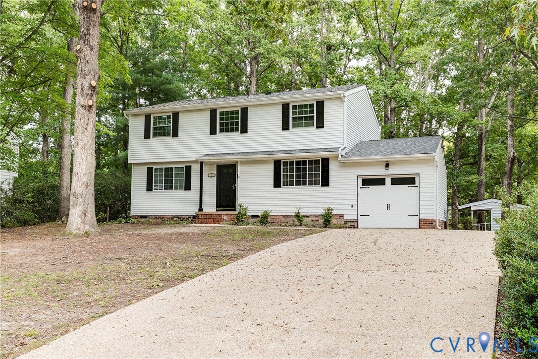 White siding exterior with black shutters, mature trees, attached single-car garage, and a gravel driveway