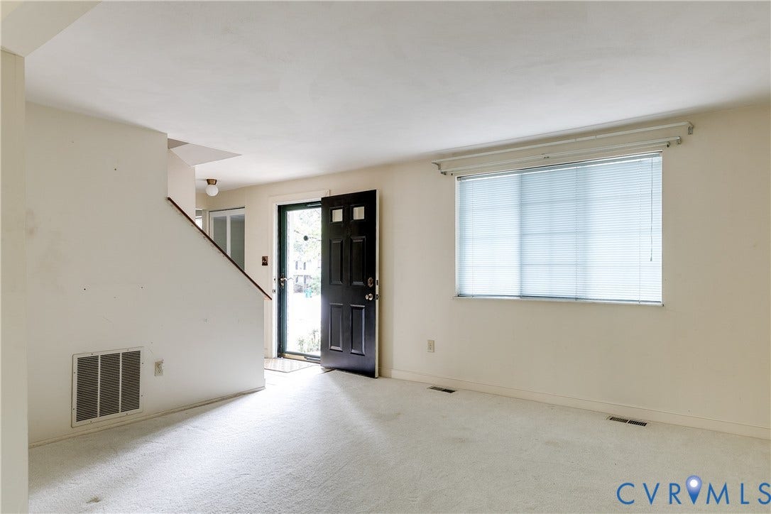 Wall-to-wall carpeting in a neutral tone, a large window with horizontal blinds, a dark-paneled entry door, and a staircase with a wood-finish handrail