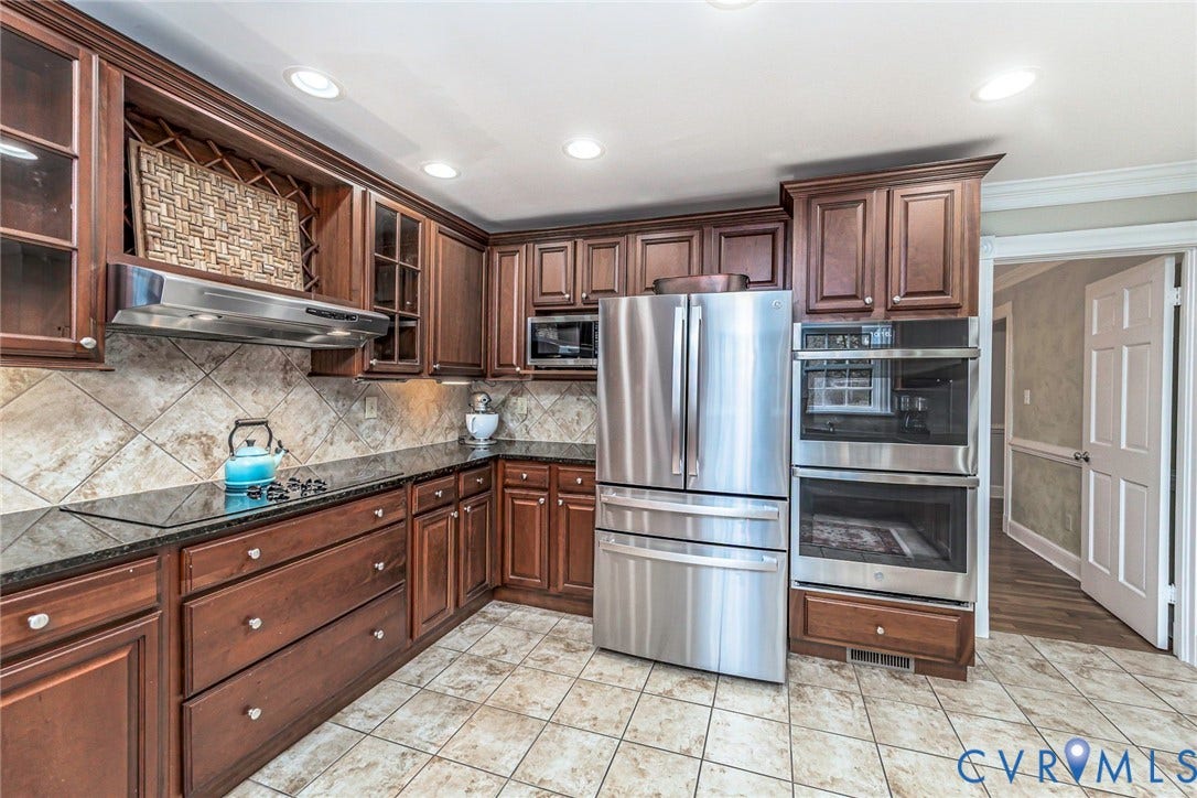 wood cabinetry with glass-front accents, stainless steel appliances, black granite countertops, tile flooring, and a diagonal tile backsplash