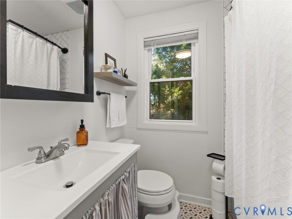 3933 Alma Avenue, Richmond, VA 23222 Single vanity with an integrated sink and brushed nickel faucet, a frameless wall mirror, subway tile shower surround, and a window with a roller shade
