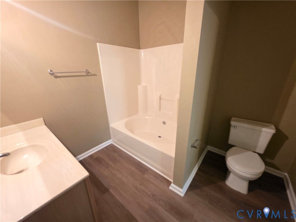 Wood-finish flooring with white baseboards, a built-in bathtub with white surround, a toilet, and a vanity with an integrated sink