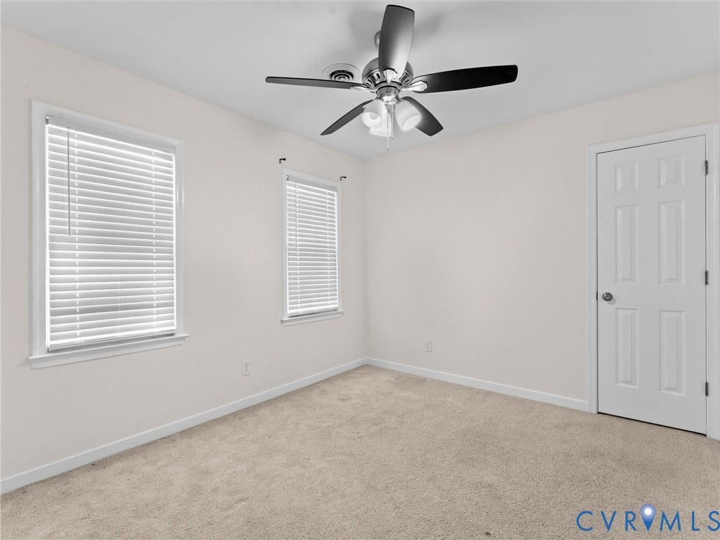 Plush carpet flooring, neutral wall tones, two windows with blinds, a ceiling fan with integrated lighting, and a paneled door with a round knob