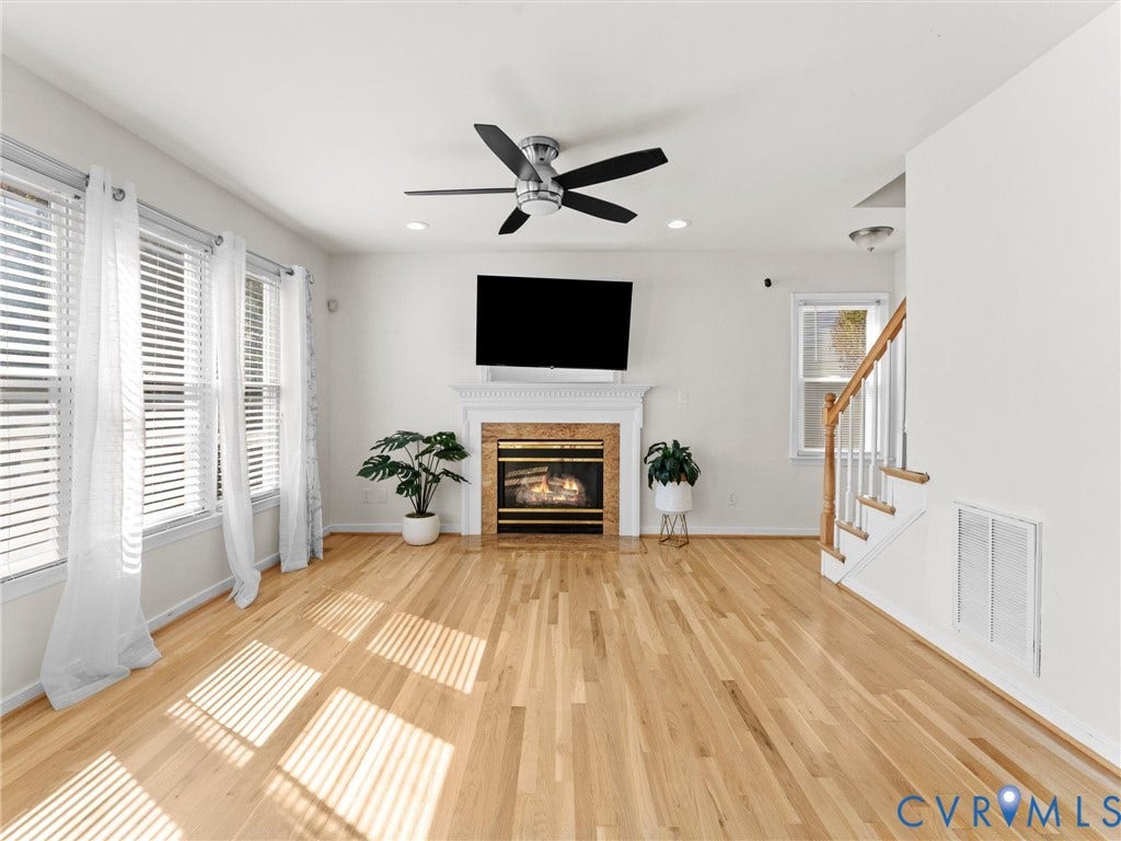 Hardwood flooring throughout, a fireplace with a stone surround and white mantel, multiple windows with blinds, a ceiling fan, and recessed lighting