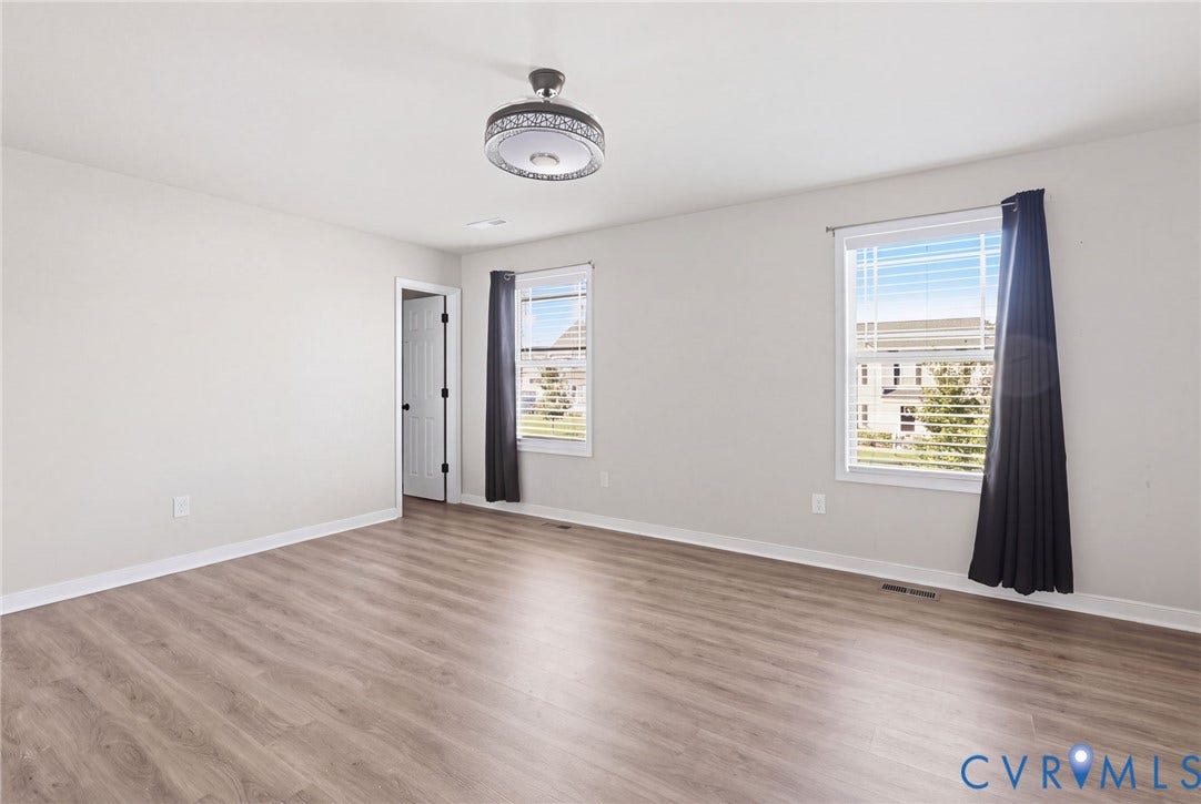 Spacious room featuring wood-finish flooring, two windows with blinds, a white paneled door, and a decorative semi-flush mount light fixture