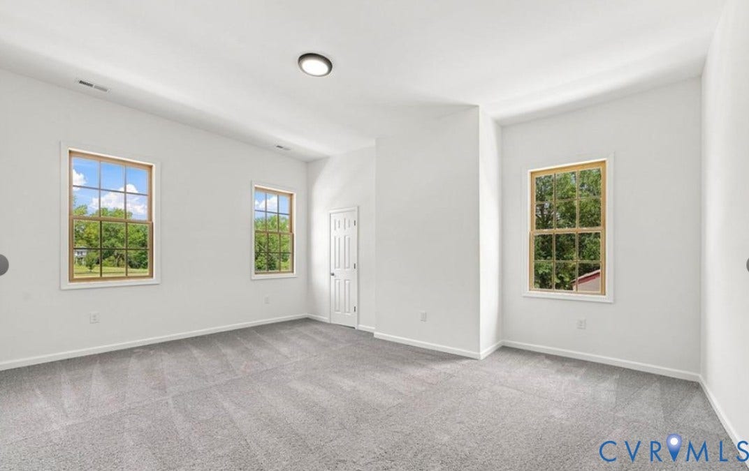 104 E Fillmore Street, Petersburg, VA 23803 Spacious room featuring light gray carpeting, neutral wall paint, two large windows with wood-tone frames, and a flush-mount ceiling light fixture