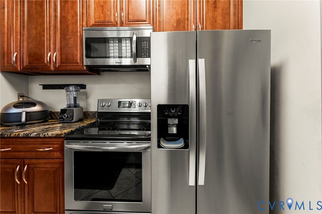 Stainless steel side-by-side refrigerator, stainless steel range with ceramic cooktop, stainless steel microwave, wood-finish cabinetry, and stone-look countertops