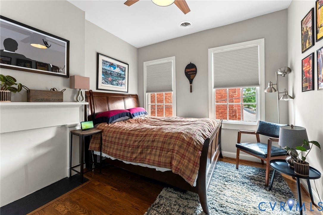 Hardwood flooring, painted fireplace surround with mantel, two windows with blinds, ceiling fan with light fixture, and light gray wall paint