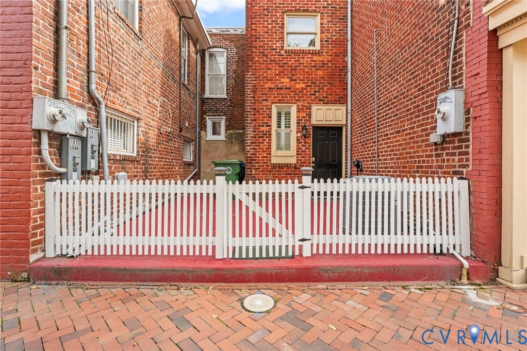 Brick paver ground surface, white picket fencing with double gates, red brick facade, and a black paneled door with a transom window