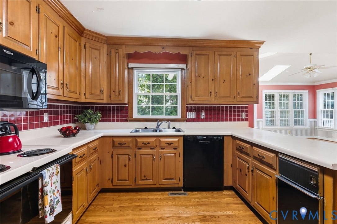 Wood-finish flooring, wood cabinetry, solid surface countertops, red tile backsplash, and a window above the sink