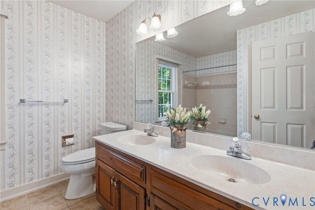 Dual undermount sinks set in a light-toned countertop, wood-finish vanity with cabinet storage, tile flooring, and a large wall-mounted mirror