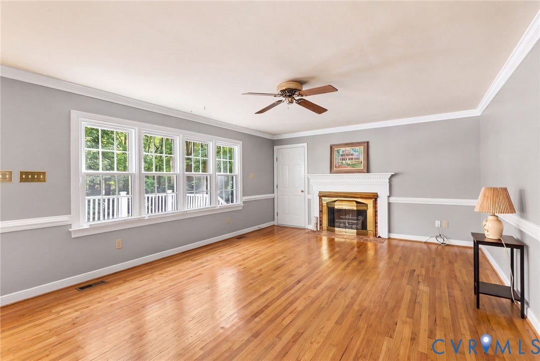 AI Version of Family Room without wall paper & carpet. Hardwood flooring, white fireplace with a gold-toned insert, multi-pane windows, ceiling fan, and crown molding