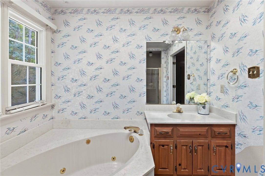 Jetted corner tub with brass fixtures, wood vanity with built-in sink, wall-mounted mirror, and overhead vanity lighting