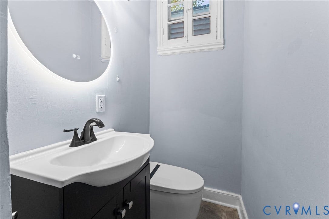 724 N 1st Street, Jackson Ward, Richmond, VA 23219 Illuminated oval mirror, white porcelain sink, dark wood-finish vanity, matte black faucet, and a window with white louvered shutters