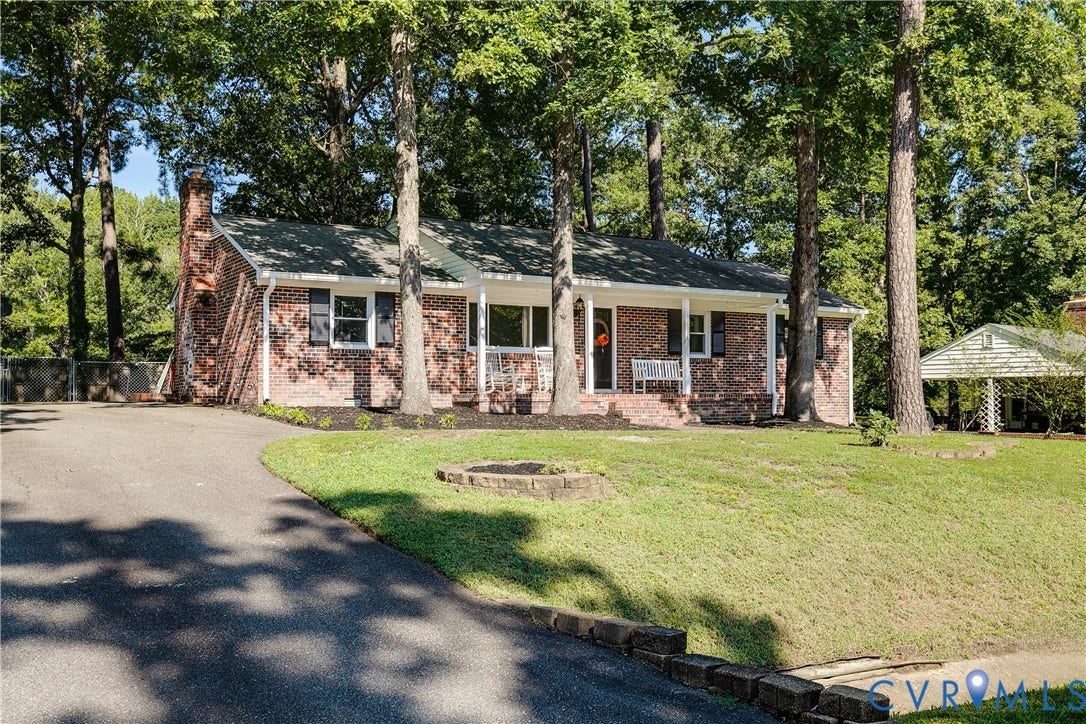 Brick exterior with a covered front porch, asphalt driveway, mature trees, and a detached carport