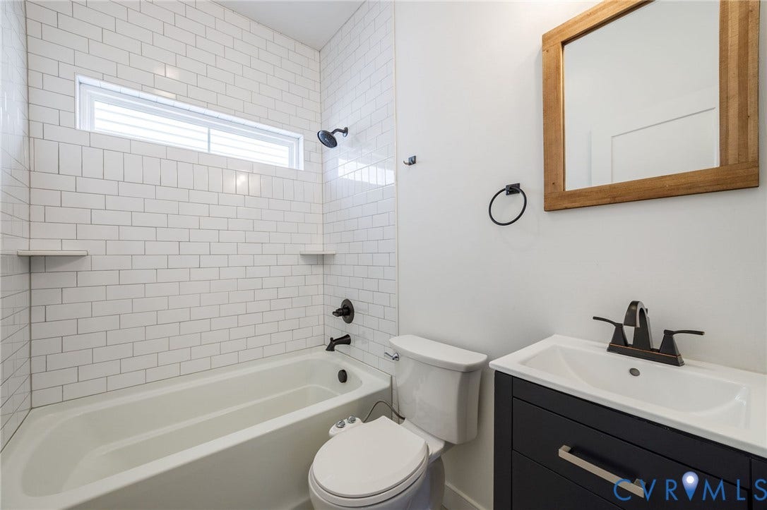 2902 Lawson Street, Richmond, VA 23224 Bathroom featuring white subway tile tub surround, dark-finish hardware, built-in shelving, a deep-toned vanity with a white basin, and a wooden-framed wall mirror