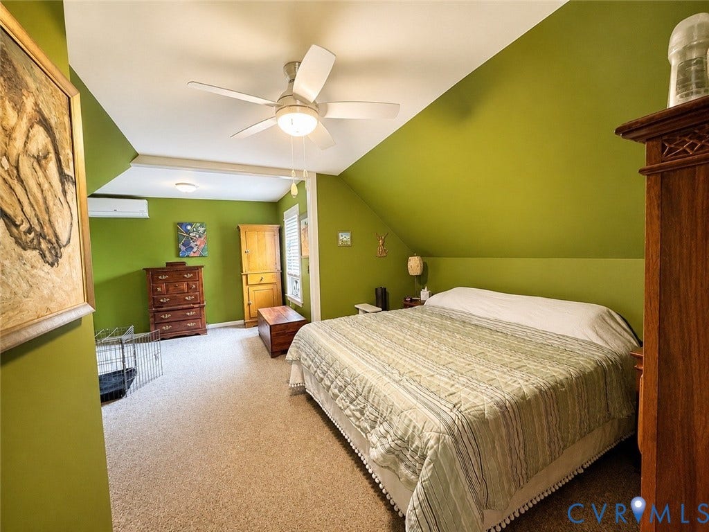 Carpeted flooring, vaulted ceiling, ceiling fan with integrated lighting, wall-mounted air conditioning unit, and window with shutters