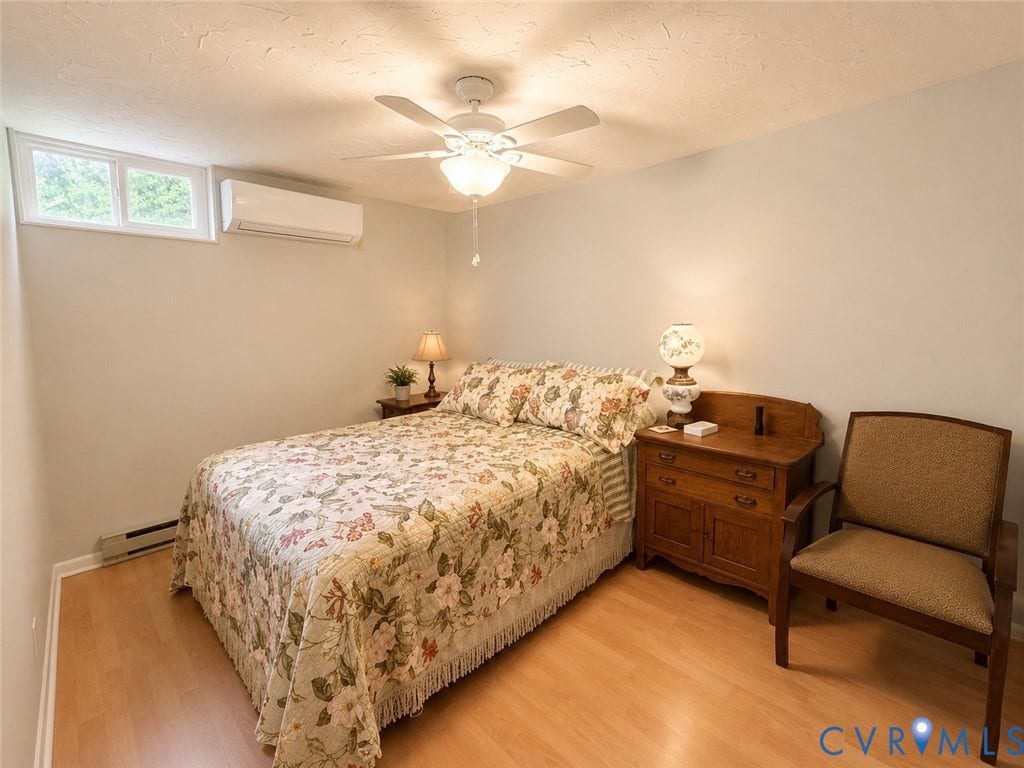 Window with white trim, wall-mounted air conditioning unit, ceiling fan with integrated lighting, wood-finish flooring, and baseboard heating