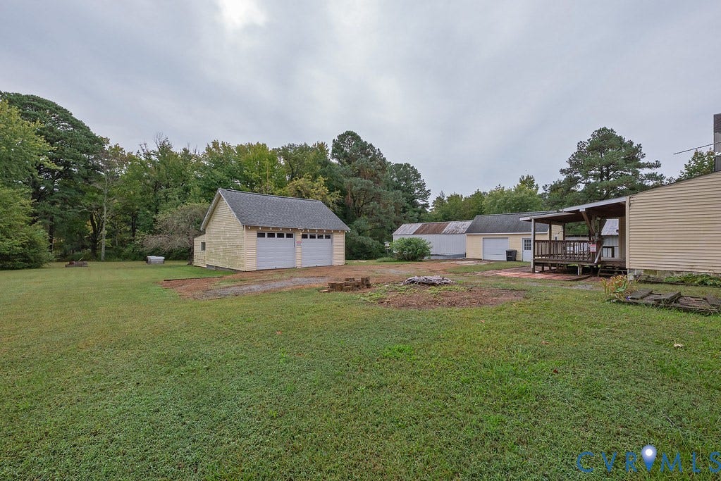Expansive grassy lot featuring mature trees, a detached two-car garage, multiple outbuildings, and a covered wood-deck patio