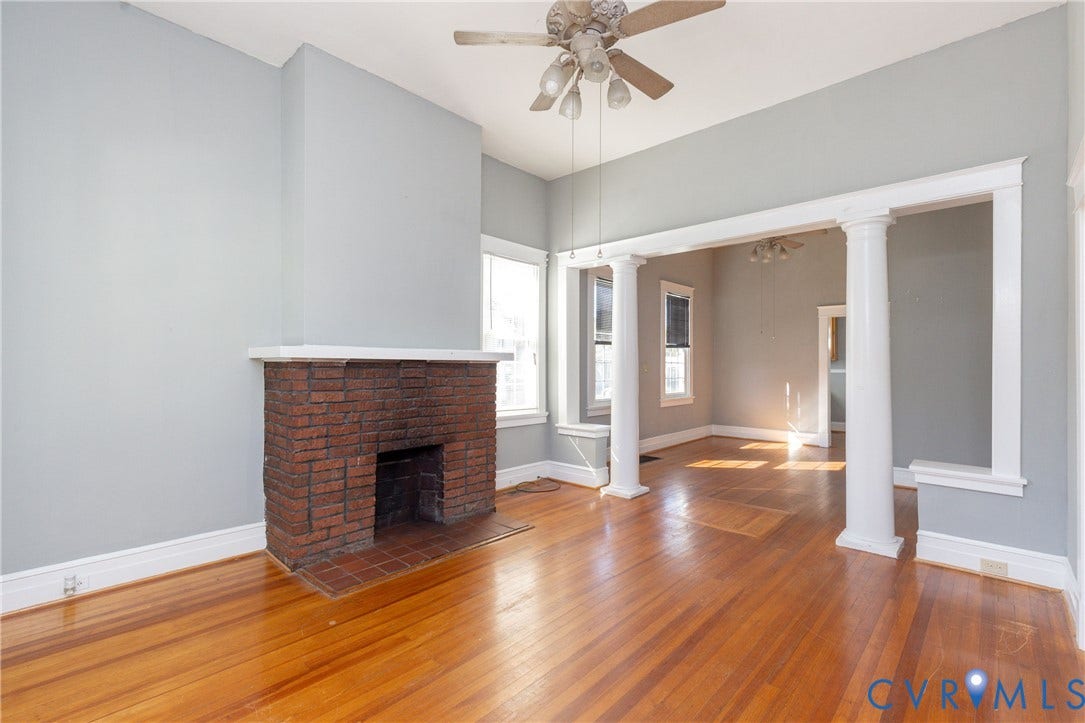 Interior room featuring wood-finish flooring, a red brick fireplace with a mantle, decorative columns, and a ceiling fan with integrated lighting