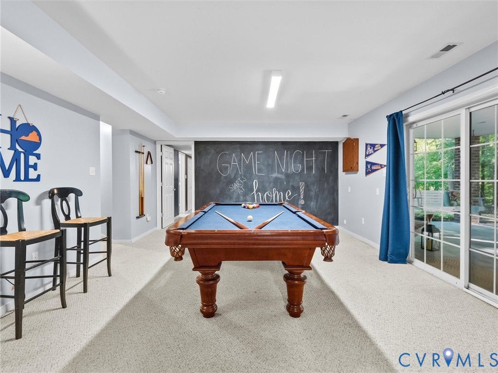 Finished recreation room featuring a regulation pool table, neutral carpeting throughout, recessed and linear ceiling lighting, large glass sliding doors with blue curtain panels, and chalkboard wall accent with "Game Night at Home" script